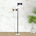 Three-arm floor lamp with black, white, and brass drum shades against marble wall with potted tropical plant on wood floor