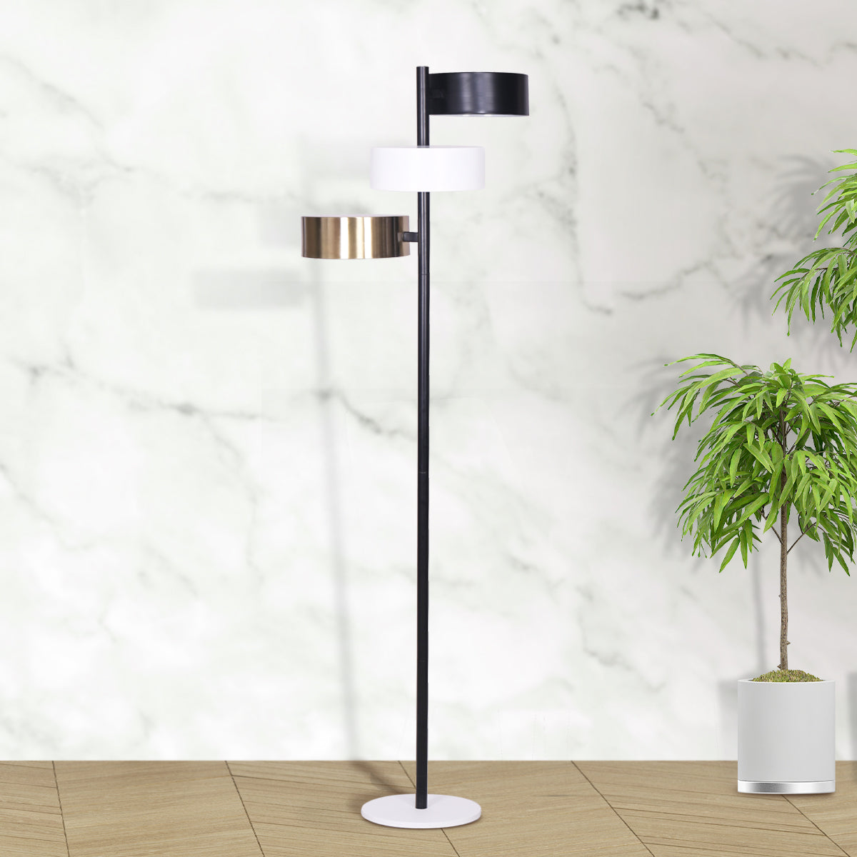 Three-arm floor lamp with black, white, and brass drum shades against marble wall with potted tropical plant on wood floor