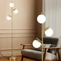 Gold 3-globe floor lamp with three white globe shades next to grey armchair in a beige paneled room, detail close-up on right