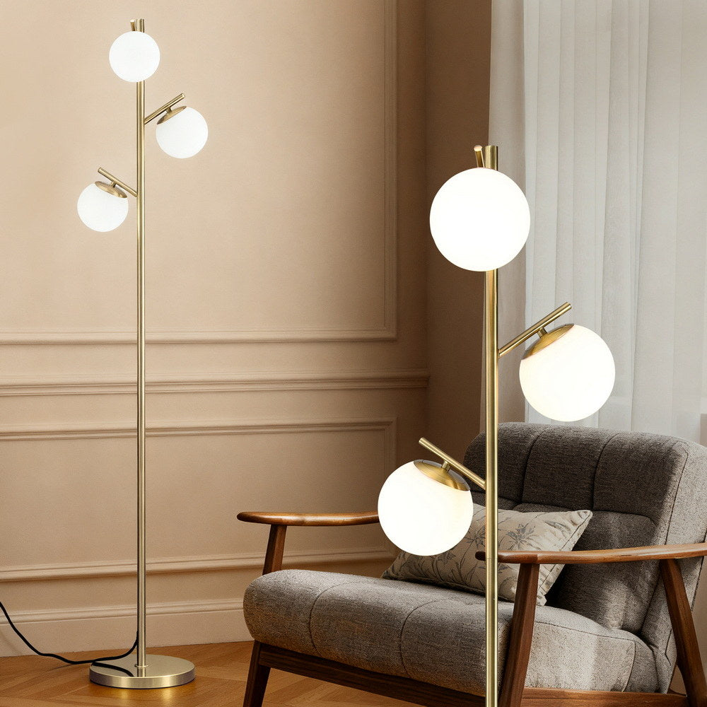 Gold 3-globe floor lamp with three white globe shades next to grey armchair in a beige paneled room, detail close-up on right