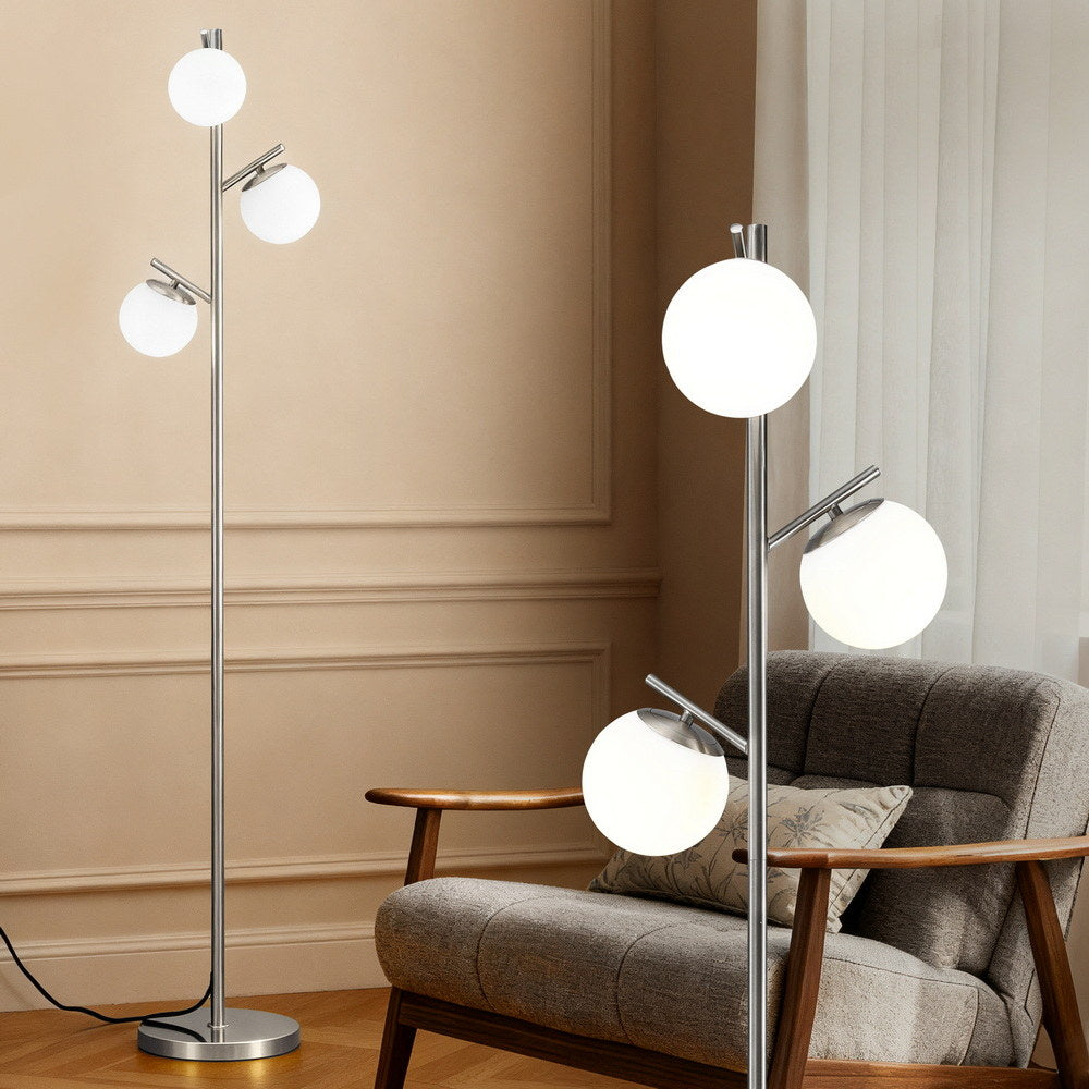 Silver 3-globe floor lamp with three white globe shades next to grey armchair in a beige paneled room, detail view on right