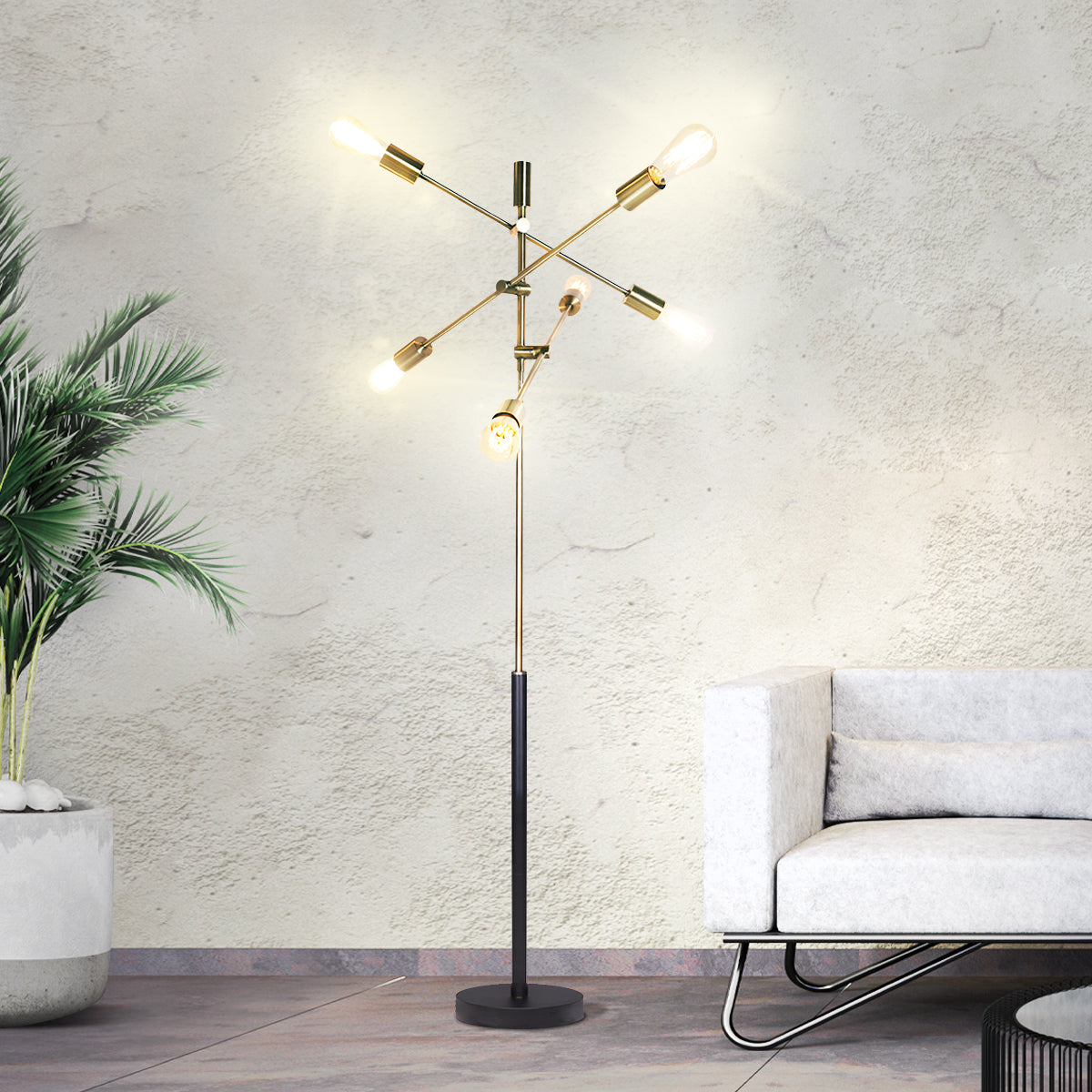 Five-arm sputnik floor lamp with lit Edison bulbs and gold brass arms beside white sofa and palm plant against textured wall