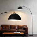 Arc floor lamp with lit black dome shade and brass fitting arching over a brown leather sofa, black marble base visible