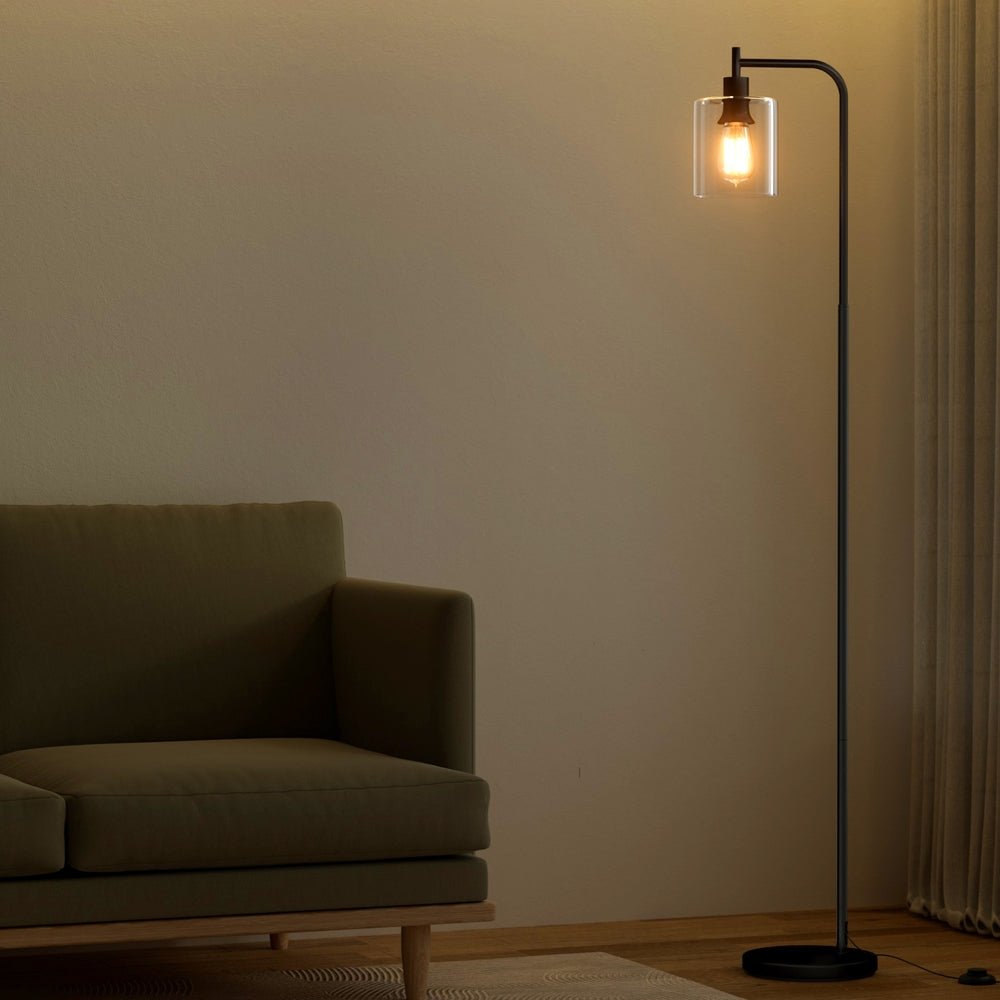 Black arc floor lamp with glowing filament bulb in clear glass shade beside dark green sofa in dim living room