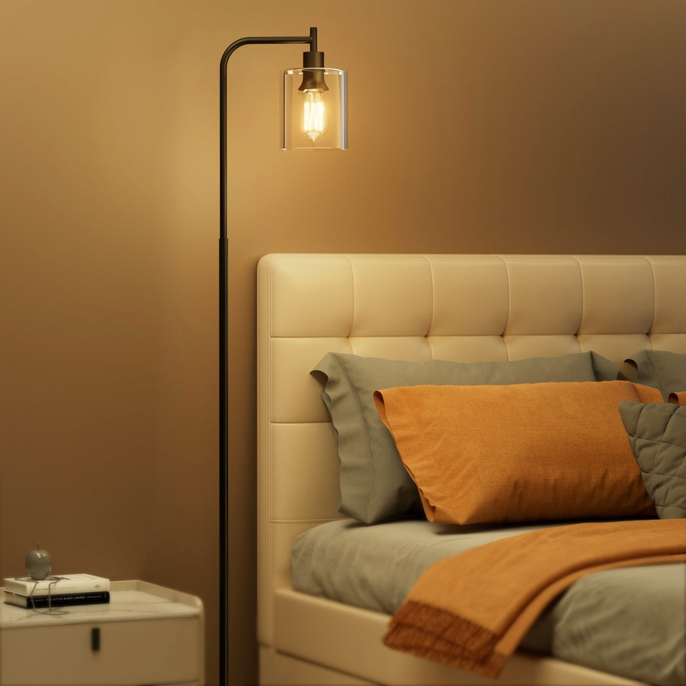 Black arc floor lamp with glowing filament bulb in clear glass shade beside tufted bed with orange and grey pillows