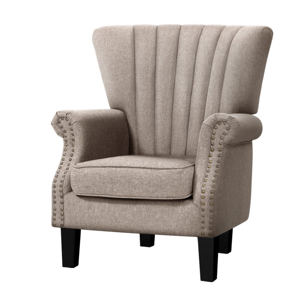 Beige fabric channel-stitched wingback armchair with nailhead trim on arms, angle view on white background