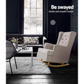 Beige fabric tufted wingback rocking chair in dark living room with dark sofa, coffee table and large window, text: Be swayed