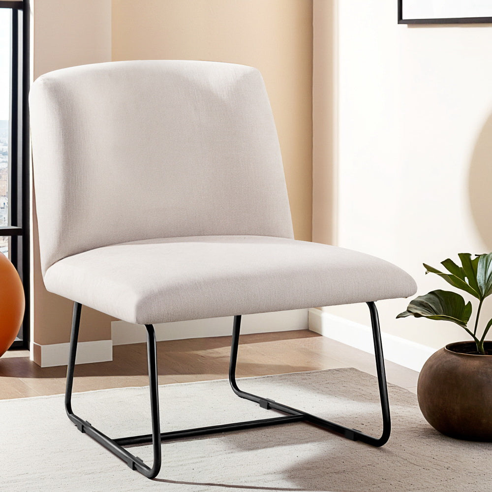 Beige linen armless accent chair with black sled base on cream rug in warm-toned room with plant and city window view