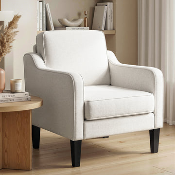 Beige linen armchair with structured back and piped edges, round wood side table with candle, bookshelf background