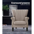 Beige fabric channel-stitched nailhead wingback armchair in dark lifestyle room with plant, text: Enamoured presence