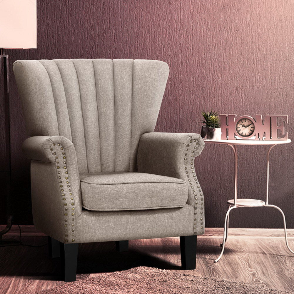 Beige fabric nailhead wingback armchair beside white side table with clock and HOME letters, mauve textured wall background