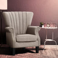 Beige fabric nailhead wingback armchair in mauve room with white side table, clock and HOME letters, angle view