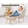 Beige fabric wingback armchair and footstool in use with person seated feet on ottoman and Enamoured presence text