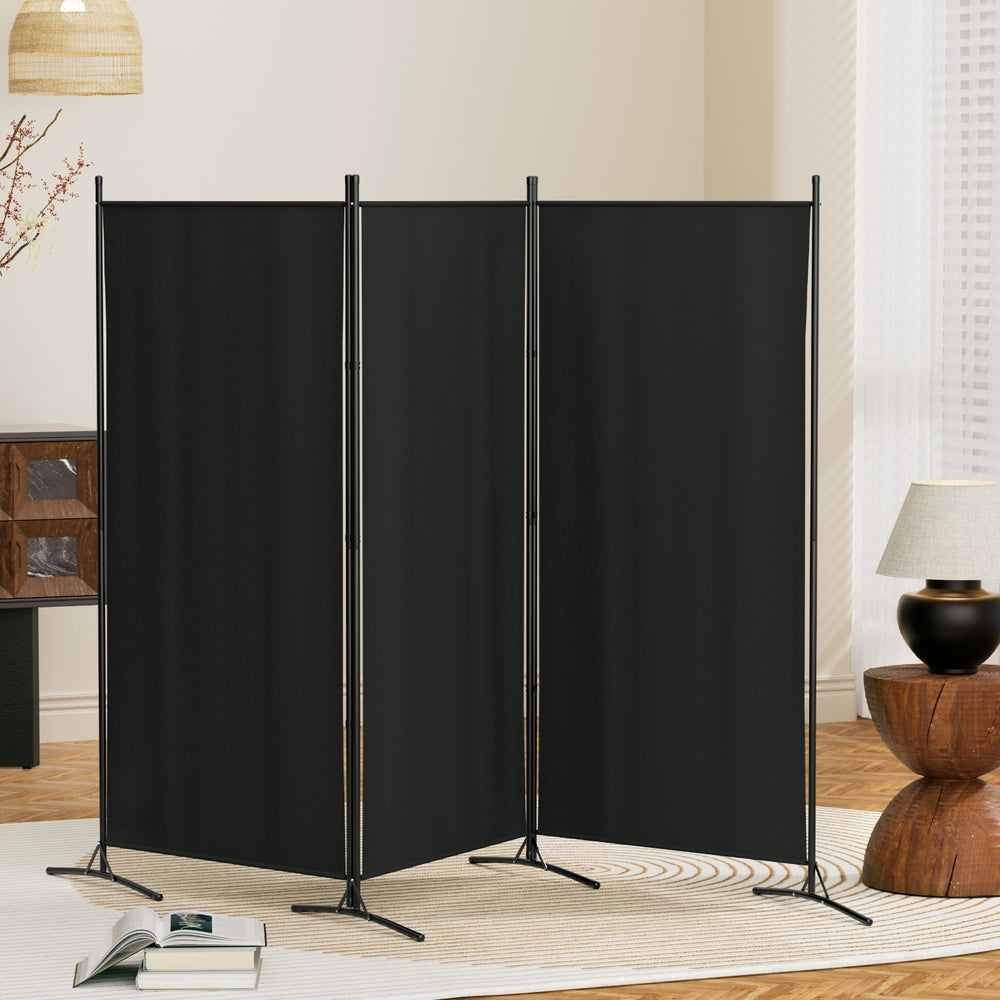Black 3-panel fabric room divider zigzag configuration in living room lifestyle with blossoming branch lamp and open book