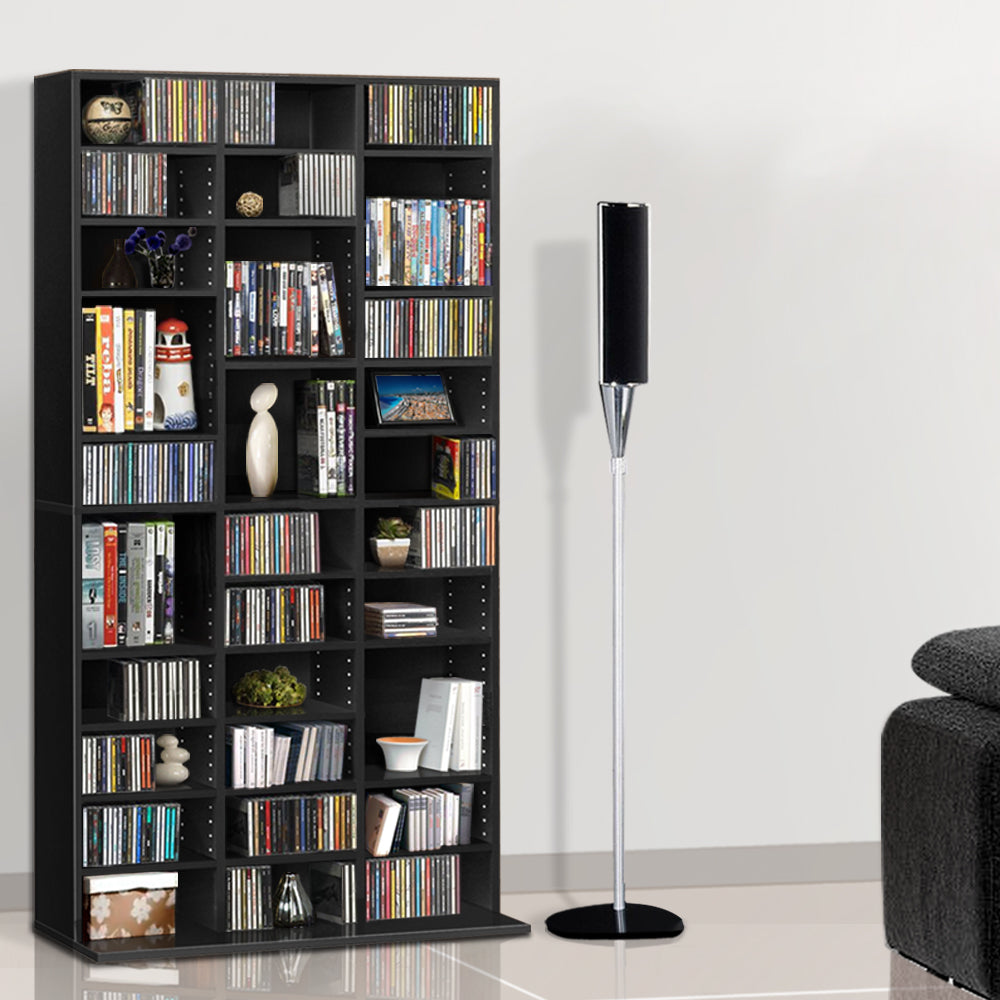 Black multi-column CD and DVD media storage shelf loaded with discs in living room beside floor speaker