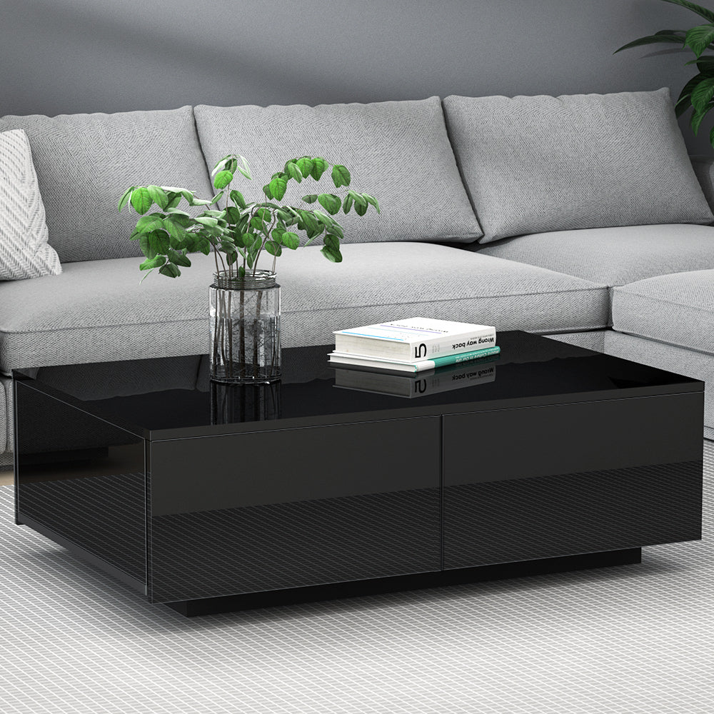 Black high-gloss rectangular coffee table with four drawers and textured panel front, styled with a plant and books in a