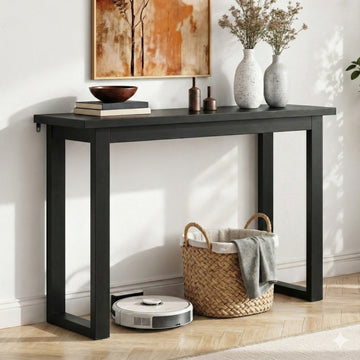 Black console table with open U-shaped legs in hallway, vases on top, wicker basket and robot vacuum underneath