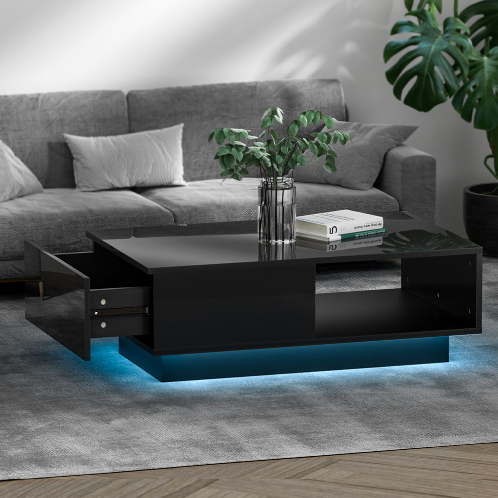 Black high-gloss LED coffee table in living room, drawer pulled open, blue LED base, plant vase and books on top, grey sofa