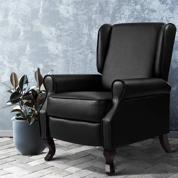 Black faux leather wingback recliner armchair with rolled arms beside rubber plant on brick floor, blue-grey textured wall