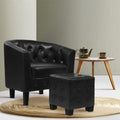 Black PU leather tub chair and ottoman on round rattan rug beside wooden side table with teapot, sage green wall, front angle