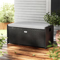 Black rattan outdoor storage bench with cream cushion on wooden deck, white weatherboard wall and plant in background