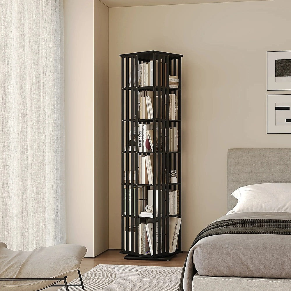 Black 6-tier rotating tower bookshelf with slatted panels filled with books in bedroom beside grey upholstered bed