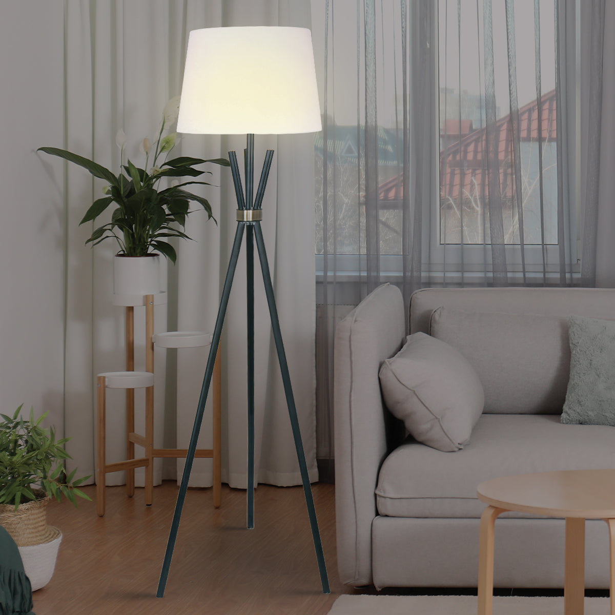 Floor lamp with lit white tapered shade in living room, dark green tripod legs beside beige sofa, wooden plant stand behind