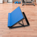 Blue adjustable calf stretching board on wooden floor in home gym with dumbbells and exercise equipment in background