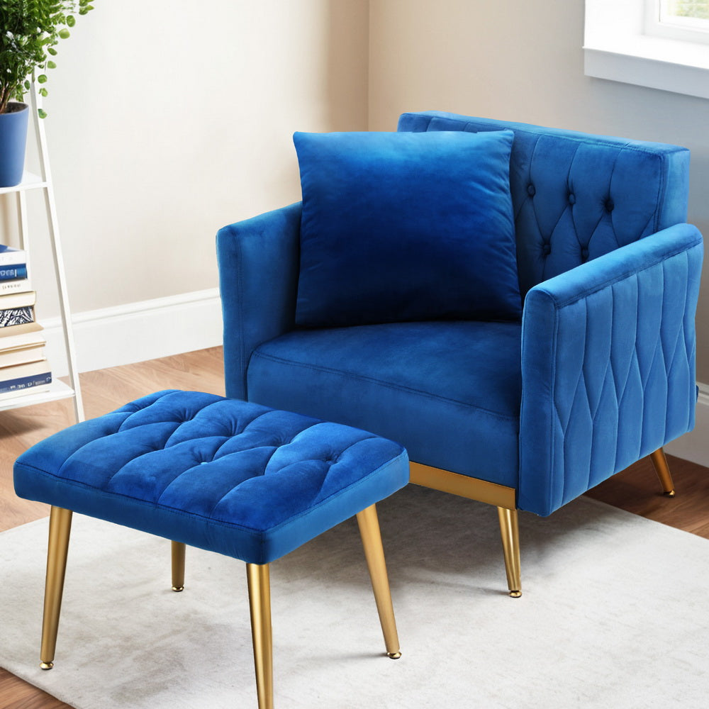 Blue velvet armchair with cushion and tufted ottoman in room setting with bookshelf, cream rug, window and gold legs