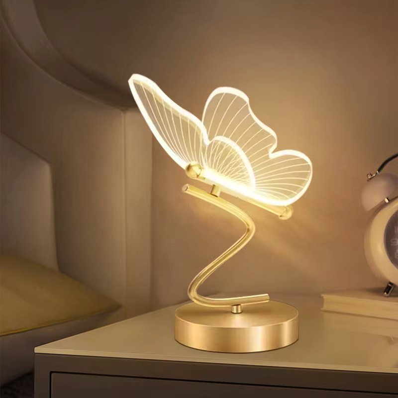 LED table lamp with acrylic butterfly-shaped illuminated shade on gold S-curve stem on bedside table beside alarm clock