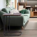 C-frame side table with walnut-look top beside green sofa holding white illustrated mug, bookshelf in background