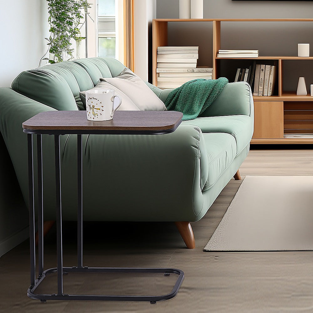 C-frame side table with walnut-look top beside green sofa holding white illustrated mug, bookshelf in background