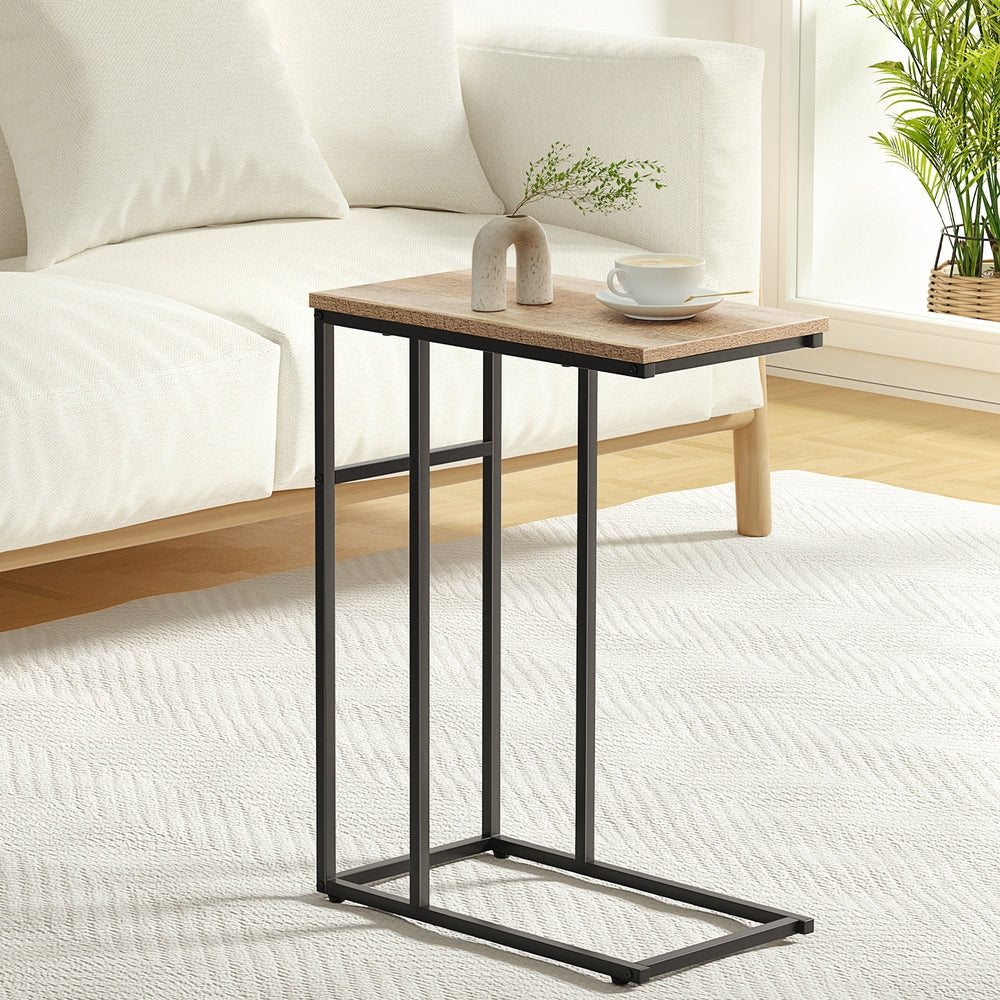 C-shaped side table styled next to a white sofa with a cup and small vase on top, on a white rug in a bright living room.