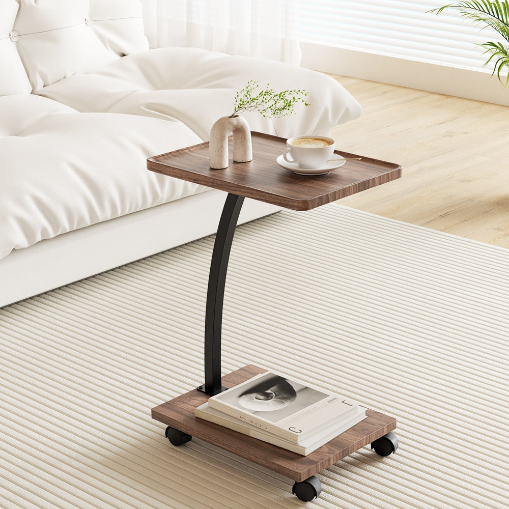 C-shaped mobile side table on castors with walnut wood tray top and lower shelf, black curved frame, beside white bed