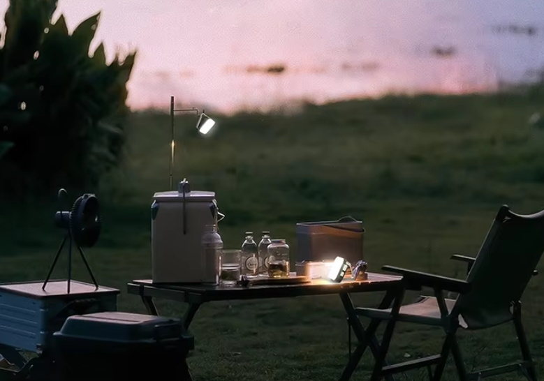Camping setup at dusk on grass by water, folding table with cooler, jars, fan and compact light, chairs nearby