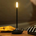 Black candle-style LED table lamp with thin stem and small rectangular light head, warm glow on wooden surface