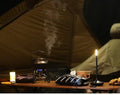 Black candle-style LED lamp on camping table inside tent with steaming kettle on camp stove, mug and walnuts