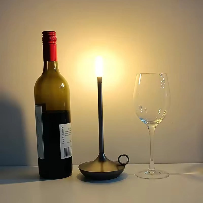 Black candle-style LED lamp between wine bottle and empty wine glass on white surface, warm ambient light