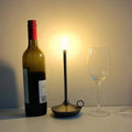 Black candle-style LED lamp between wine bottle and empty wine glass on white surface, warm ambient light