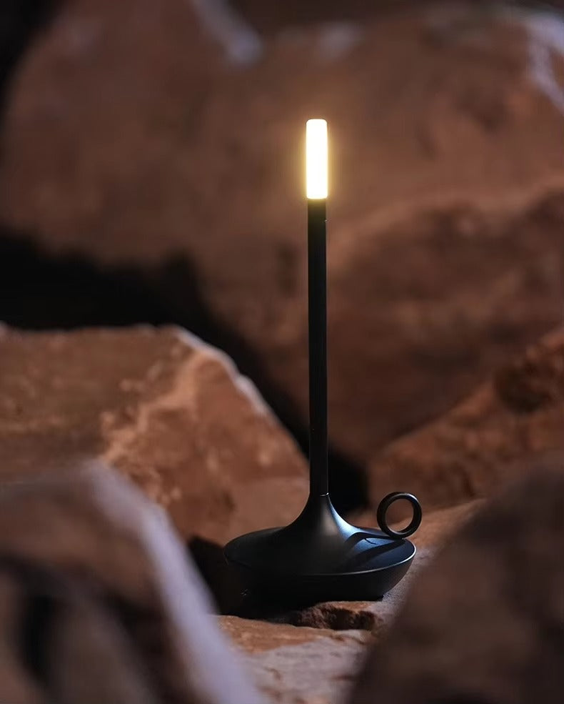 Black candle-style LED table lamp with ring handle placed among large rocks, warm white light glowing at top
