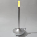 Silver candle-style LED table lamp with ring handle on round base, warm amber glow at top, grey background