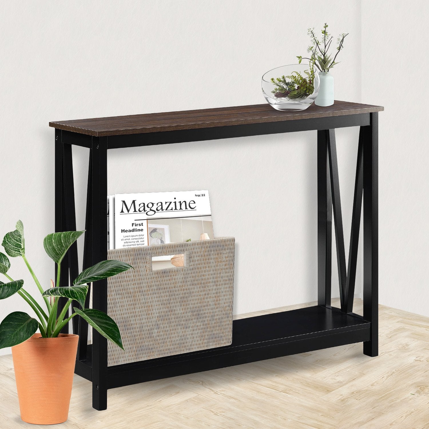 Walnut-top black X-frame console table in room, glass bowl and plant on top, wicker magazine holder on lower shelf
