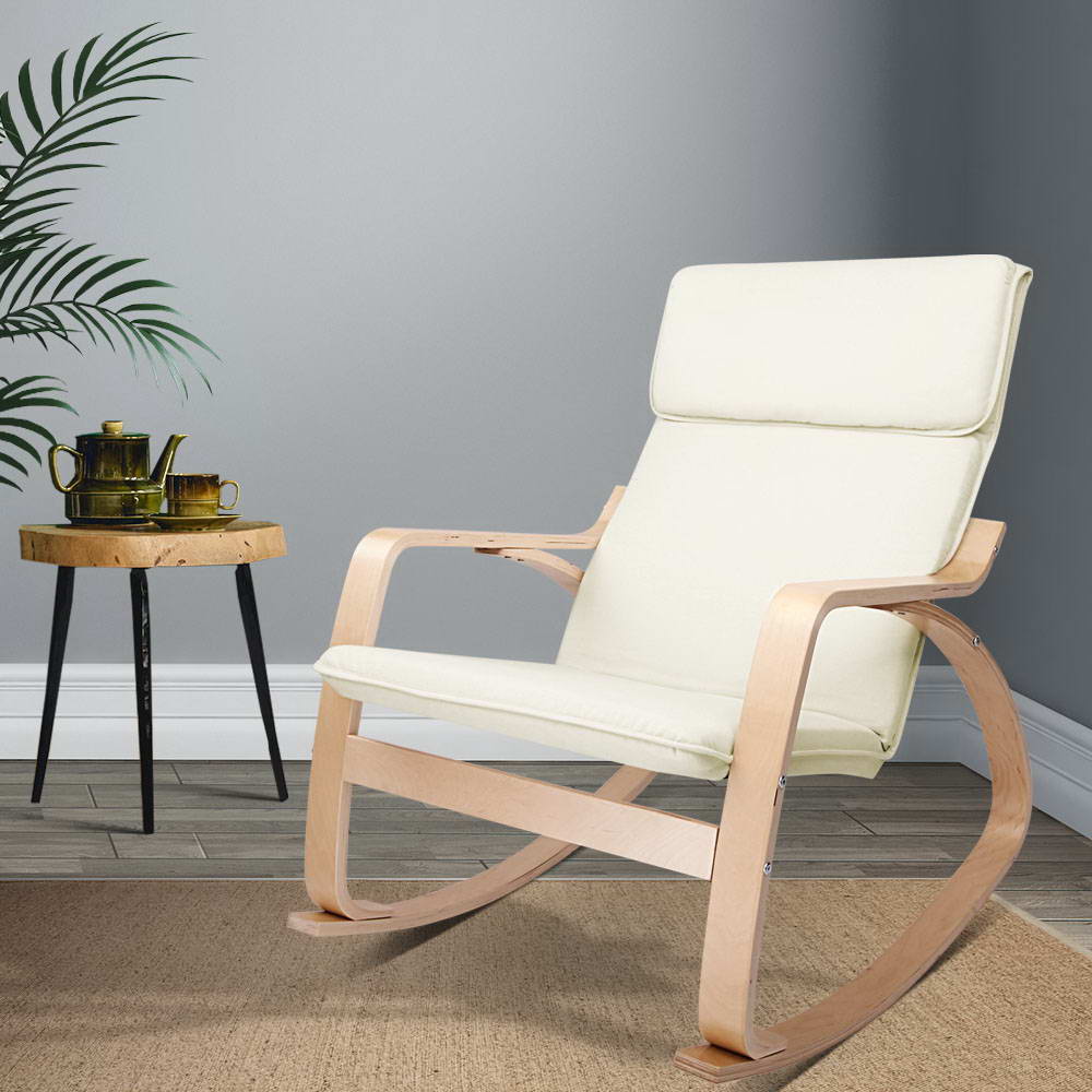 Cream fabric bentwood rocking chair beside wooden tripod stool with gold tea set, grey wall and palm plant in background