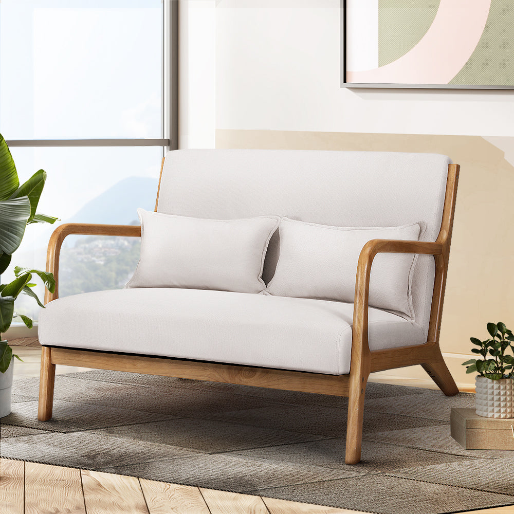 Cream fabric two-seater loveseat with solid wood curved armrests, two cushions, in living room with window and plants