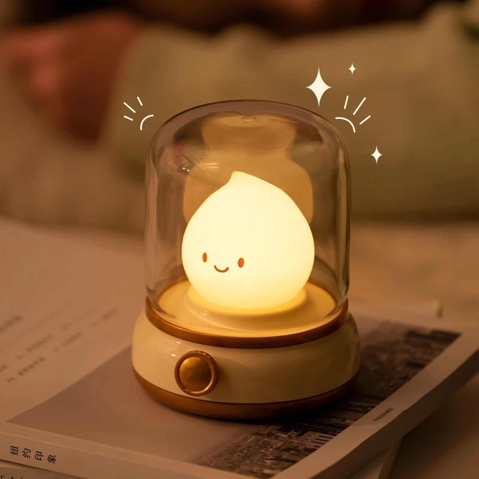 Smiley face raindrop LED lamp in glass dome with sparkle graphics sitting on top of a book on warm-lit surface