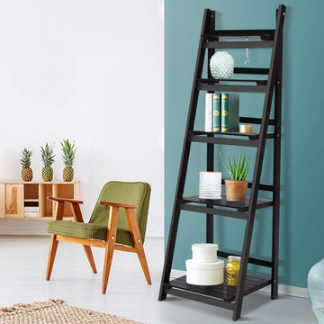 Dark espresso 5-tier A-frame ladder shelf styled with books, aloe plant and hat boxes against teal wall beside armchair