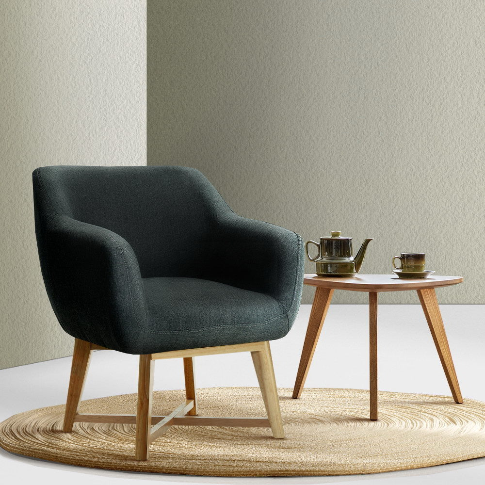 Dark charcoal fabric tub armchair on round jute rug beside wooden side table with tea set, sage green wall background