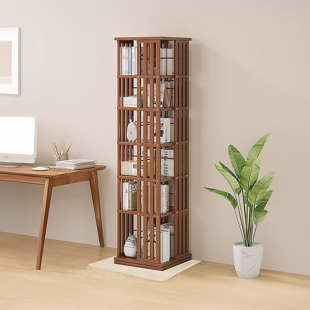 Dark wood slat rotating tower bookshelf with books in home office setting beside wooden desk with monitor and tropical plant