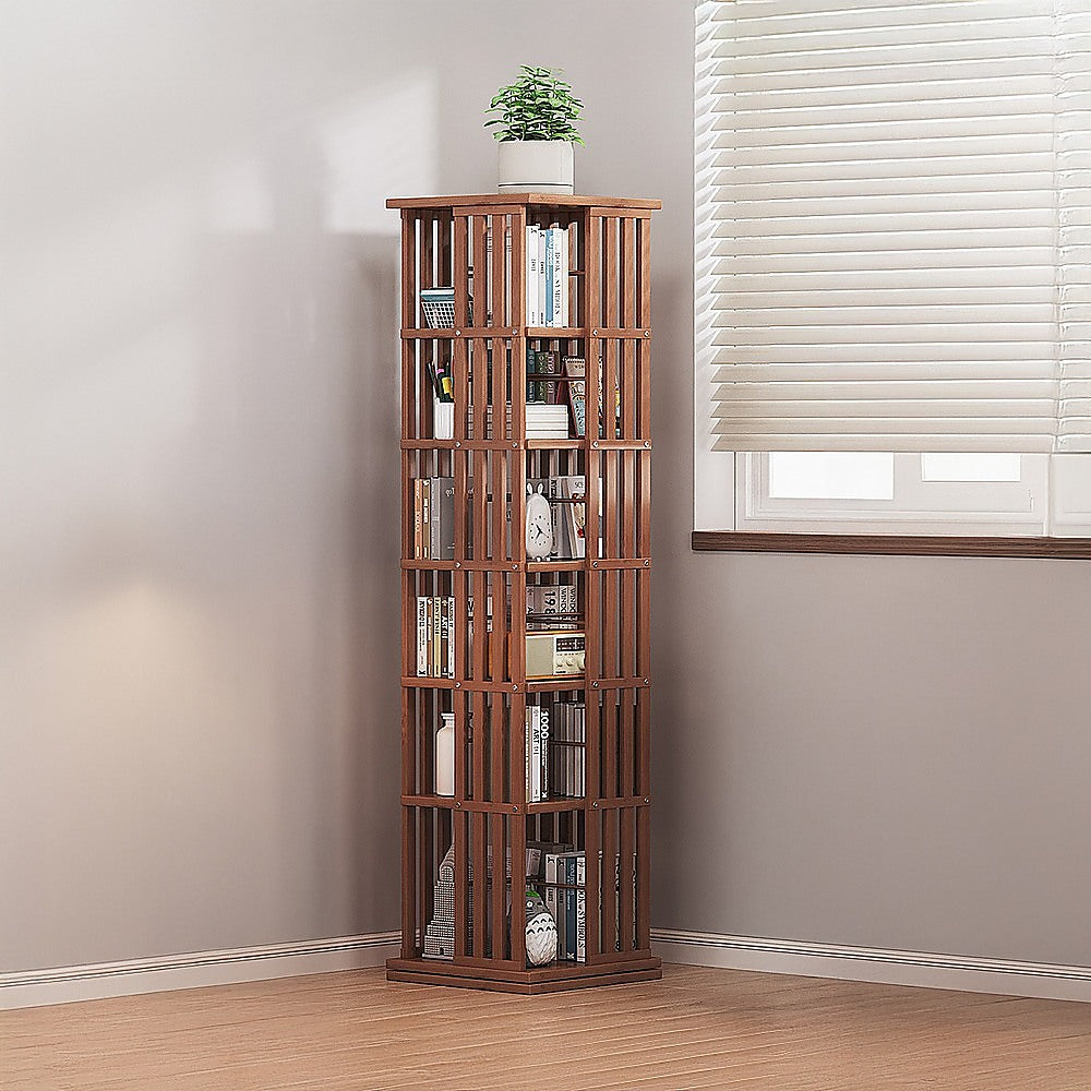Dark wood slat rotating tower bookshelf filled with books in room corner, plant pot on top, venetian blinds in background