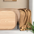 Rounded natural wood grain tray resting against a beige velvet storage ottoman with a mustard knit throw draped over one end